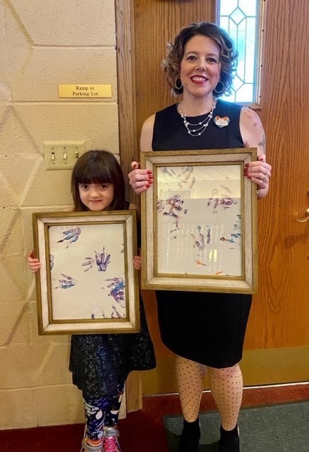 Picture of woman standing with young girl both holding framed paper covered in paint hand prints.