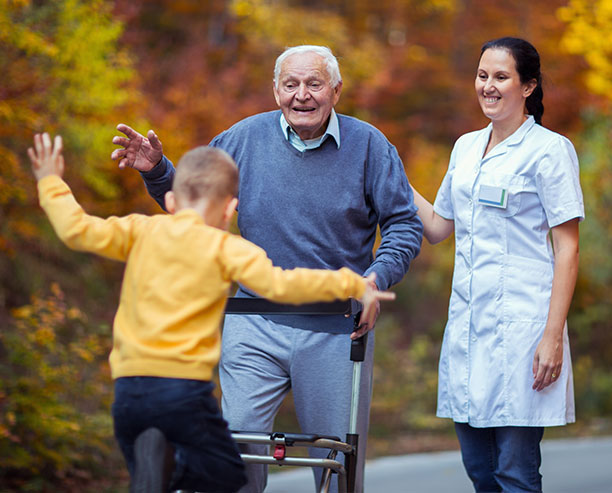 Cheerful Grandfather In Walker With Caregiver Welcoming Happy Grandson